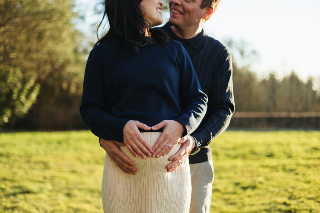 pregnant woman holding heart on belly with husband hugging from behind