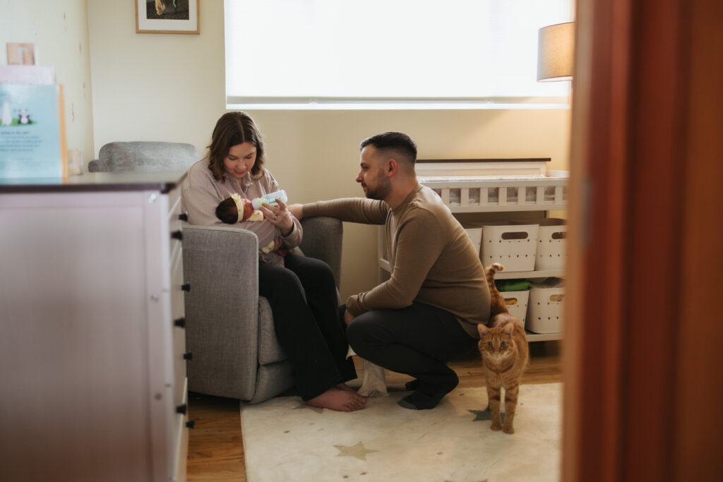 mom feeding newborn in home nursery while dad watches with orange cat