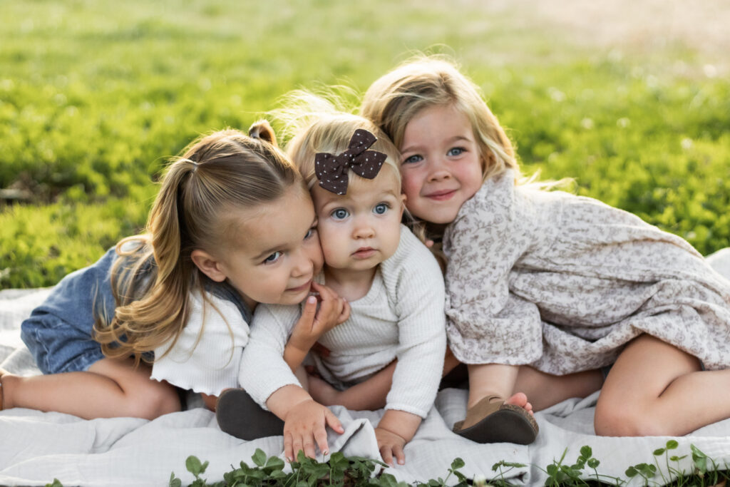 three toddler sisters hugging on a blanket in the summer on grass field.